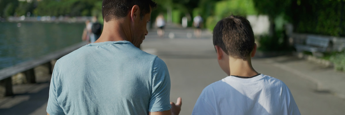 Ein Mann und ein Junge gehen nebeneinander am Wasser entlang und scheinen sich ruhig miteinander zu unterhalten.