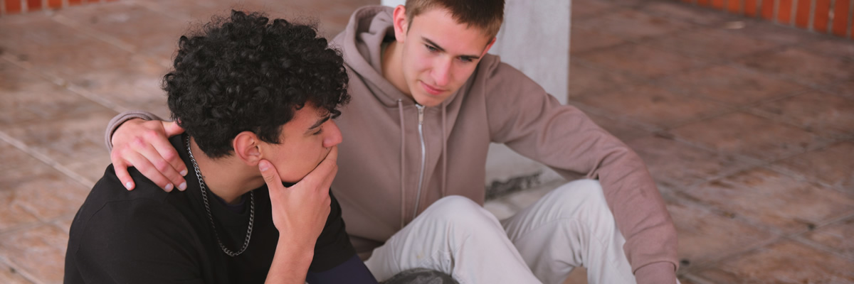 Ein Jugendlicher legt einem anderen tröstend den Arm um die Schultern, während dieser besorgt oder nachdenklich wirkt.